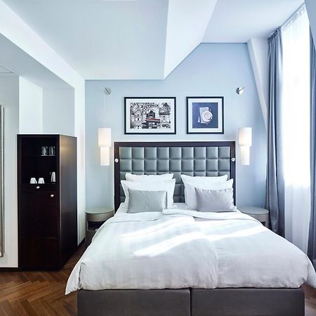 Steigenberger Hotel Herrenhof Vienna Exterior photo The photo shows a stylish hotel room with a modern design. It features a large, neatly made bed with white linens and gray accents. On the wall above the bed, there are two framed artworks. The room has large windows with light gray curtains, allowin