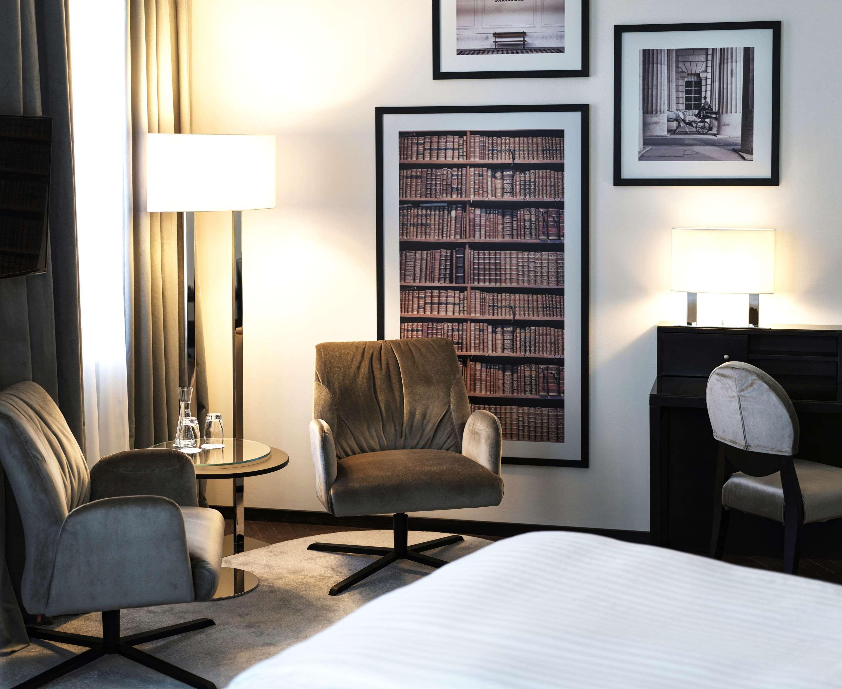 Steigenberger Hotel Herrenhof Vienna Exterior photo The photo shows a modern hotel room or lounge area. It features two stylish chairs, a small round glass table, and a standing lamp adding warm light to the space. Behind the chairs, there is a large framed print that resembles a bookshelf filled with