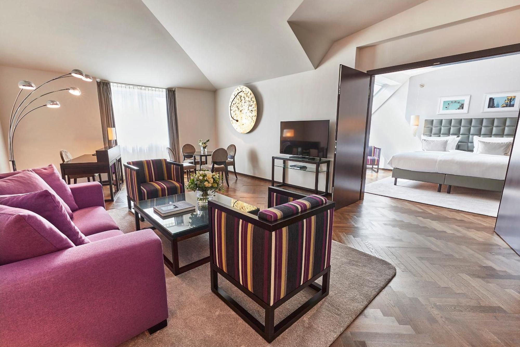 Steigenberger Hotel Herrenhof Vienna Exterior photo The photo shows a modern, stylish living space that appears to be part of a hotel suite. The room features a cozy seating area with a purple sofa and two striped chairs around a glass coffee table. A flat-screen TV is mounted on the wall. Large windo